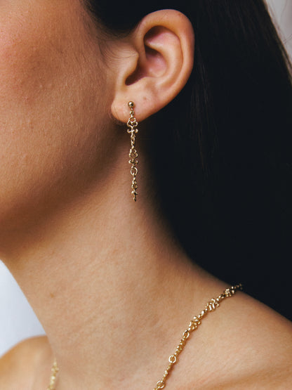Close-up of a wonan wearing handmade gold long earrings and a necklace against a dark background