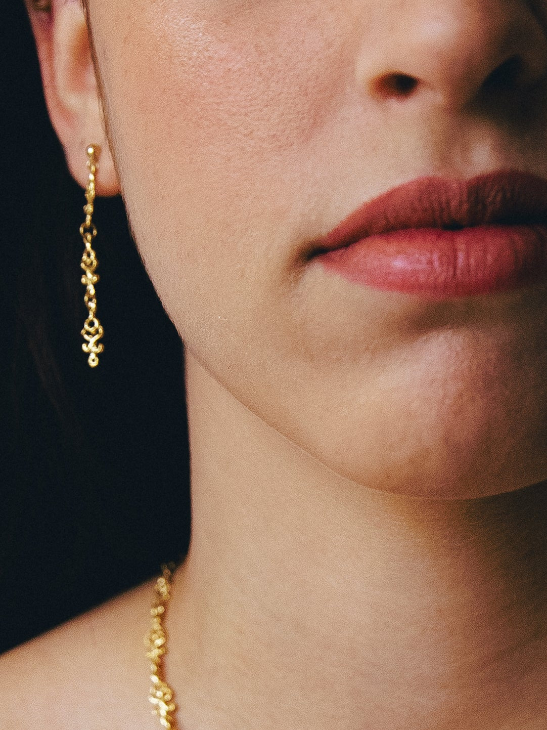 Close-up of a person wearing a gold necklace and earrings against a dark background