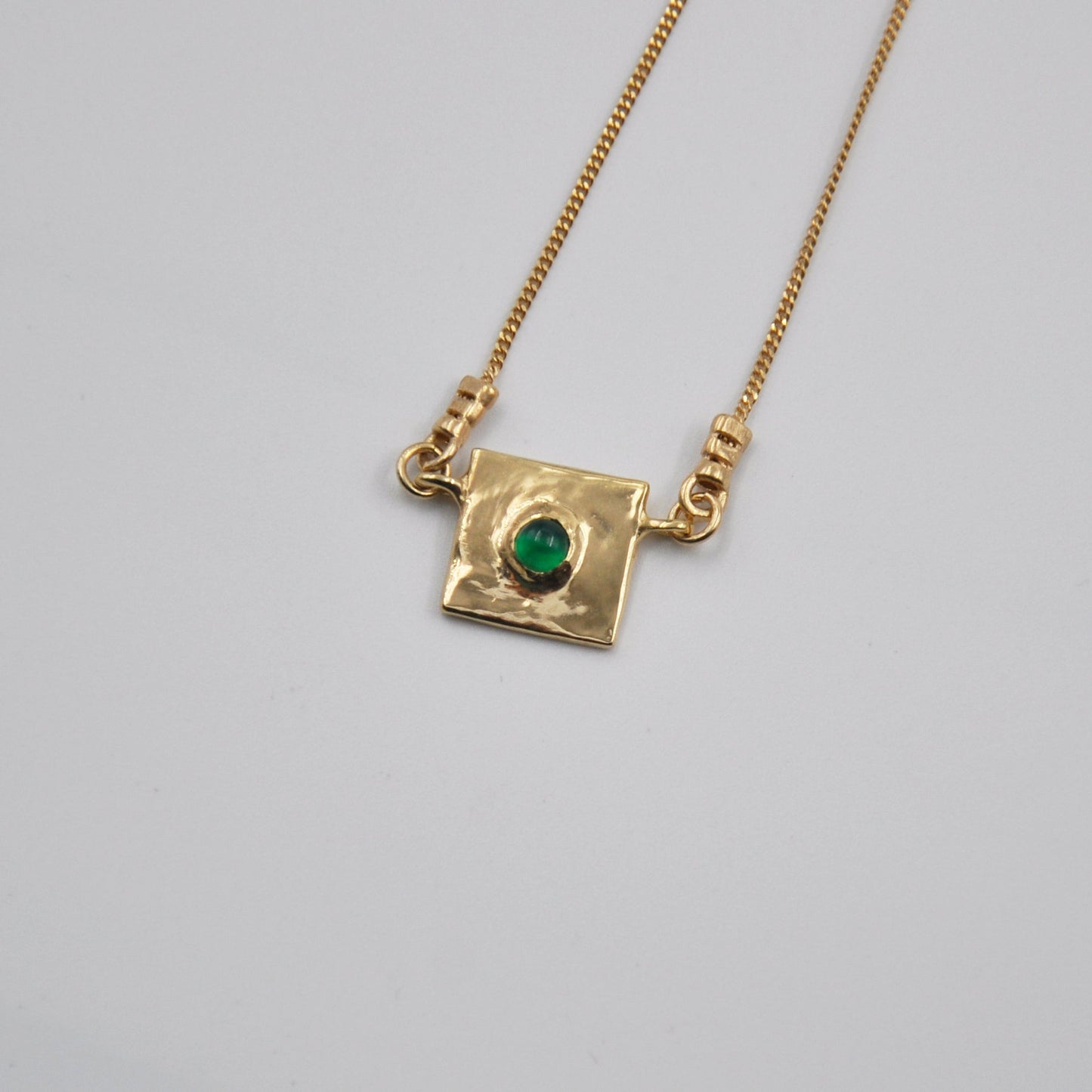 Gold necklace with a square pendant featuring a green gemstone on a light gray background