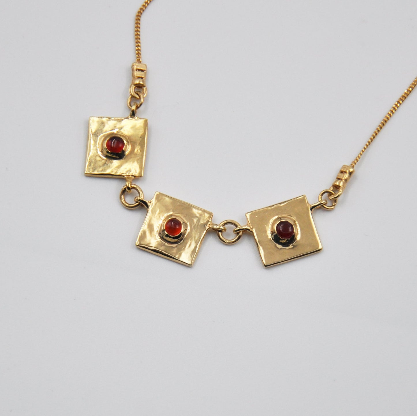 Gold necklace with square pendants and red stones on a light gray background