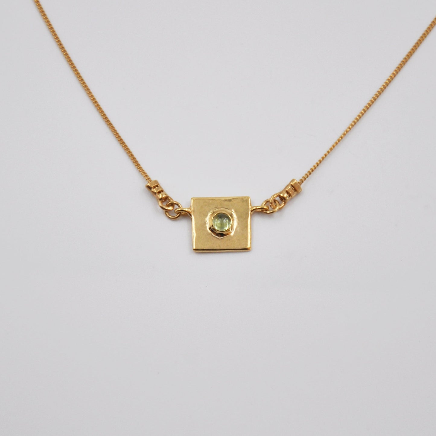 Gold necklace with a square pendant featuring a green gemstone on a light gray background